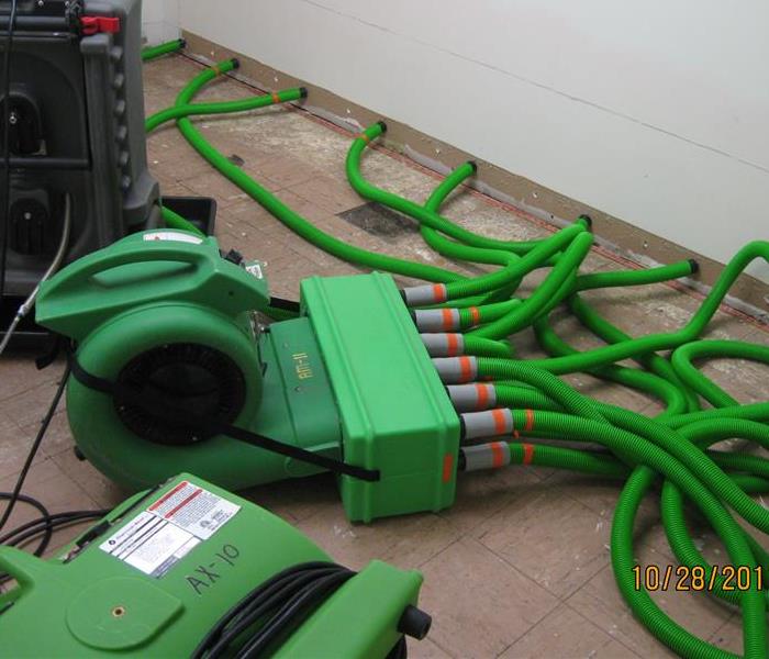 green SERVPRO equipment on floor with hoses inserted into sheetrock